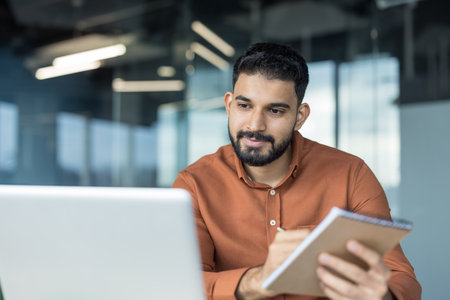 Young man concentrating on his work, looking at a laptop screen and writing in a notebook in a modern office environment, symbolizing focus and digital learningの写真素材