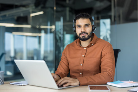 Young indian customer service professional wearing headset, focused at laptop in modern office, providing tech support and helpdesk assistance for remote corporate clientsの写真素材