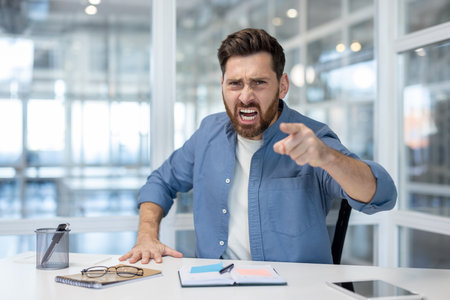Angry man screaming and pointing a finger sitting at a desk in a modern office, expressing frustration, aggression, stress, and dissatisfaction in a workplace environmentの写真素材