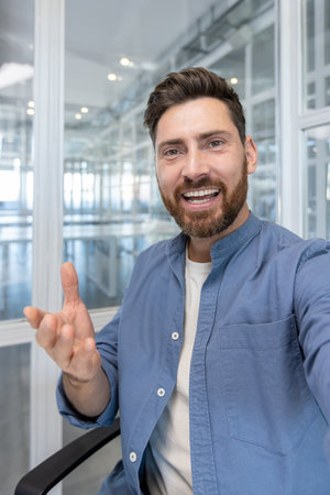 Businessman looking at camera in a video call, smiling and gesturing, communicating from a modern office environment, representing remote work and online connectionの写真素材