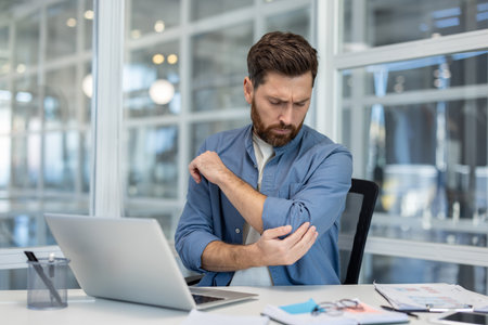 Man experiencing discomfort and pain in his elbow, touching the injured joint while sitting at a modern office desk, highlighting common work-related health problems and overuse injuriesの写真素材