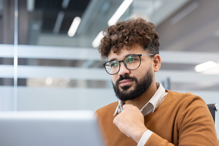 Young businessman with glasses and curly hair stares thoughtfully out the window in a modern office,...