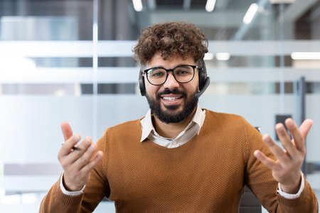 Smiling bearded man working as customer service in an office, wearing a headset and glasses,...