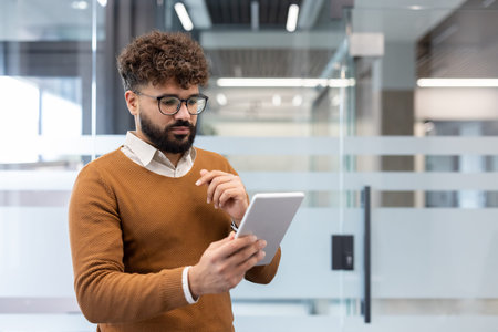 Young businessman with curly hair and glasses stands in a bright modern office, focused on a digital...