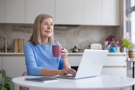 Senior woman relaxing and smiling while typing on her laptop, holding a mug of coffee, and looking away thoughtfully in her brightly lit domestic kitchen, enjoying smart work lifestyleの写真素材