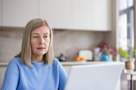 Mature woman focused on laptop in a bright modern kitchen, working remotely and browsing online,...