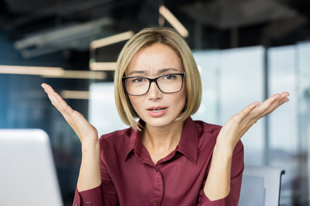 Businesswoman shrugging with open hands and a puzzled expression, indicating doubt, uncertainty, or a difficult question while working in a modern office setupの写真素材
