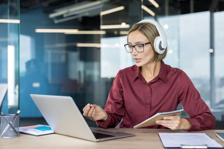 Confident businesswoman wearing headphones engages in an online meeting from a contemporary office, taking notes and discussing ideas during a professional virtual conferenceの写真素材