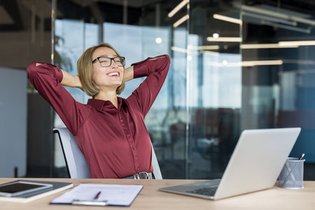 Happy businesswoman relaxing with hands behind head, smiling and laughing in a modern office, enjoying a successful break from working on her laptop on a wooden deskの写真素材