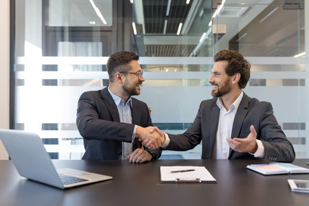 Two smiling adult businessmen in suits sitting at a table, shaking hands and making a successful agreement, partnership, and collaboration in a contemporary office settingの写真素材