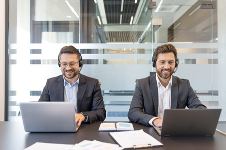 Two smiling men in suits wearing headsets and using laptops at a desk in a modern office, working as...