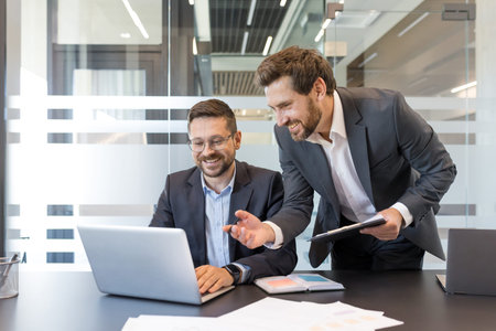 Two smiling professional businessmen are collaborating on a laptop in a bright contemporary office...