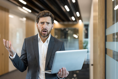 Businessman expressing confusion and frustration, raising his hand while holding a laptop, experiencing a problem or error in a modern office environmentの写真素材