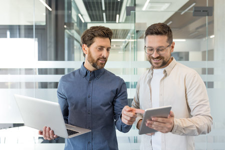 Two smiling bearded businessmen standing in a modern office, one holding a laptop while the other...