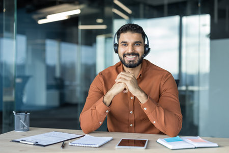 Smiling indian man wearing a headset and orange shirt sitting at his desk in a modern office,...