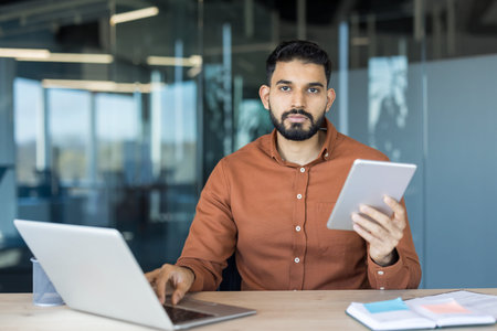 Young businessman multitasking at a modern office desk, looking at camera while working on a laptop...