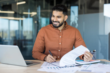Smiling indian businessman working at his desk, reviewing paperwork and using a laptop in a modern...