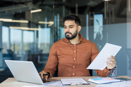 Young indian businessman in a modern office, focused on reviewing paperwork and typing on a laptop at his desk, analyzing reports and planning financial strategyの写真素材