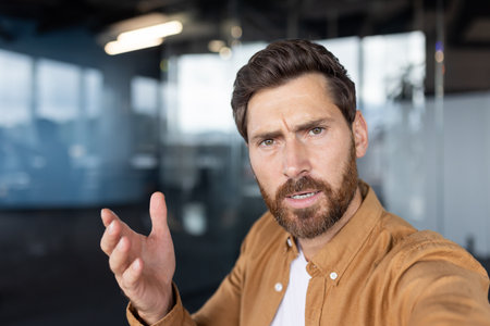 Confused man experiencing frustration and asking questions while shrugging and talking during a video call or online meeting in a modern corporate office settingの写真素材