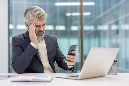 Mature businessman experiencing stress and worry while reading upsetting news on his mobile phone in...