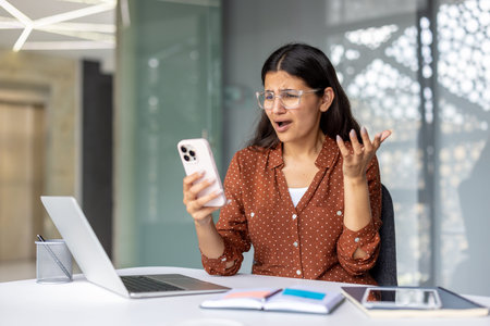 Woman reacting with frustration and stress while looking at her smartphone in an office setting,...