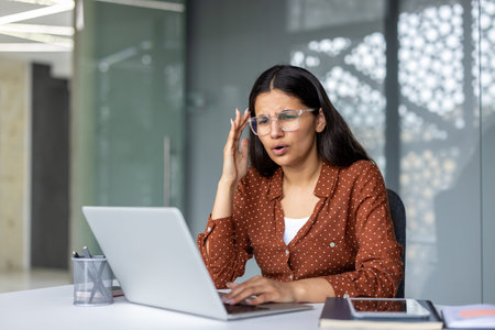 Young indian businesswoman slumped at her desk, rubbing her temples while working on a laptop,...