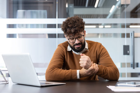 Young man sitting at his desk, suffering from wrist pain or carpal tunnel syndrome, holding his...