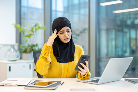 Muslim woman wearing a hijab experiencing stress and worry while reading upsetting financial or business update on her mobile phone at a modern, corporate workplace desk with laptopの写真素材
