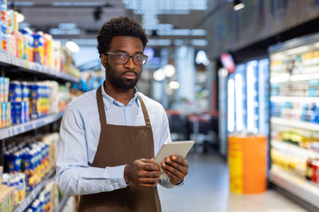African american grocery store worker wearing an apron and glasses, holding a digital tablet while...