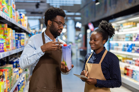 Young man and woman, both employees wearing aprons, coordinating and discussing product inventory while using a digital tablet in a grocery store aisleの写真素材
