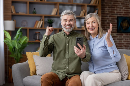 Happy senior couple sitting on a sofa, celebrating a victory or achieving success while looking at a mobile phone screen and expressing excitement and joyの写真素材