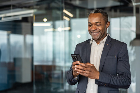 Executive man standing in a modern office building, happily using his mobile phone for professional...