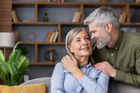Senior husband gently embracing his smiling wife in their cozy living room, sharing a warm, affectionate moment that reflects lasting love, trust and comfortable companionshipの写真素材