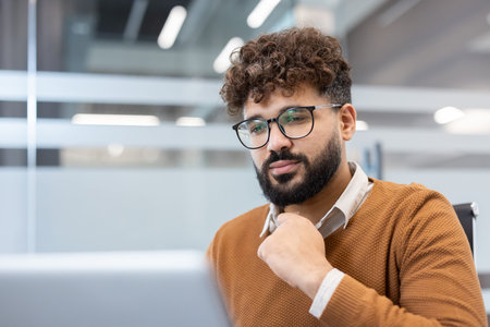 Young male employee in glasses and a brown sweater, concentrating, working on a laptop in a modern...