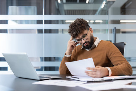 Businessman with glasses and curly hair feeling stressed and overwhelmed while reviewing documents,...