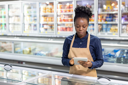 Young woman grocery store employee in an apron checks frozen food inventory and updates stock levels...