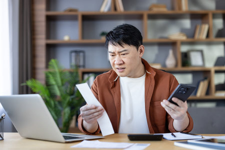 Asian man sitting at a home office desk, holding a receipt and smartphone, looking worried while...