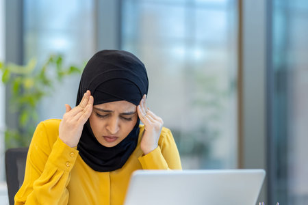 Muslim businesswoman is experiencing a painful headache and stress while working on her laptop in an modern office, feeling overwhelmed and needing to manage the symptomsの写真素材