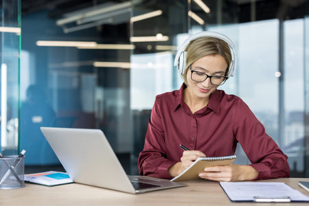 Young woman wearing headphones and glasses, watching webinar on laptop, writing notes in a notebook,...