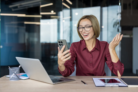Businesswoman laughing on a smartphone video call, enjoying a remote collaboration from her modern...