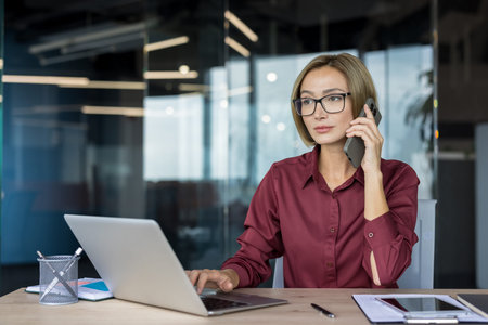Confident businesswoman talking on a smartphone while simultaneously typing on a laptop,...