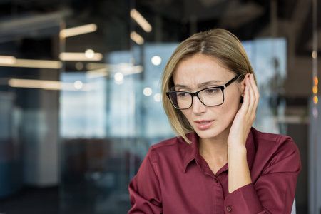 Adult businesswoman in glasses cupping ear, struggling to hear or understand a conversation in a modern office, depicting concepts of hearing difficulty, confusion, and communication problemsの写真素材