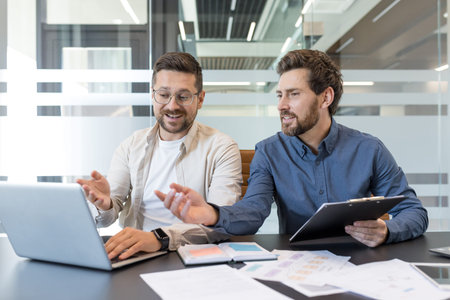 Two businessmen discussing project details and ideas while working together on a laptop, fostering...
