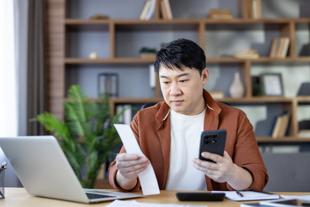 Asian man analyzing a paper receipt and using a modern smartphone for tracking expenses, managing...