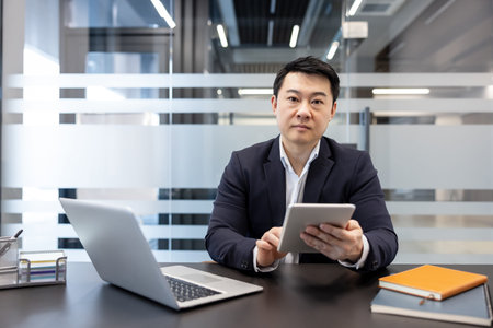 Asian businessman in a contemporary office, seated at a desk using a digital tablet with laptop and...