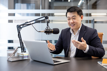 Asian man speaking into a professional microphone and looking at a laptop, broadcasting content in a...