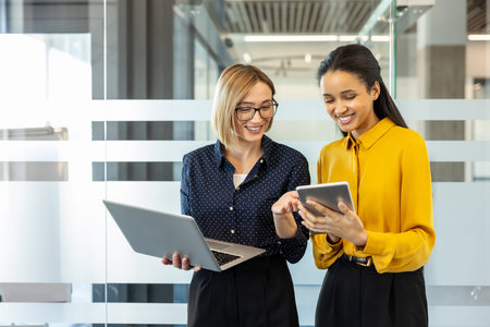 Two smiling businesswomen standing in an office, actively collaborating and discussing work using a...