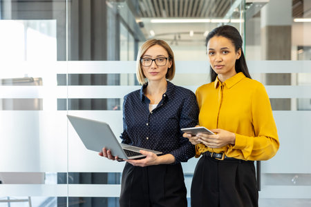 Two confident businesswomen, one holding a laptop and the other a tablet, standing together in a...