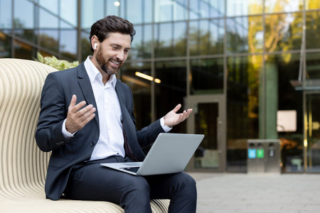 Caucasian businessman in a suit sitting on a modern bench, using a laptop and wireless earphones for a video conference or remote work outside a contemporary office buildingの写真素材