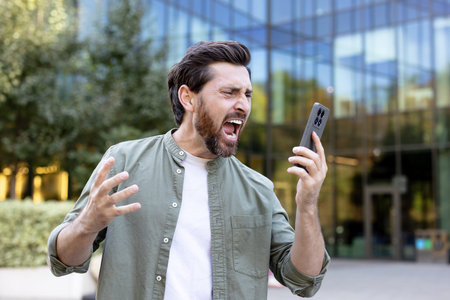 Bearded man shouting at his smartphone outdoors, expressing anger, frustration and stress from...